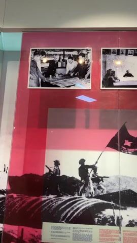 The video pans across a museum display case. The display features several black and white photographs mounted on a vibrant pink background. At the top, two smaller photographs show groups of people in formal settings, one appearing to be a meeting or planning session. Below these, a large, dramatic photograph depicts a group of figures silhouetted against a landscape, one of whom is raising a flag with a star on it. In front of this large photograph are informational text panels. At the bottom of the display, two more black and white photographs are visible, one showing people in a somewhat cramped, possibly underground space, and another depicting a gathering around a table. The entire display is behind glass, and reflections are visible on the surface.