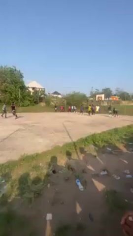 The video shows an outdoor basketball court in a daylight scene. Several individuals are present. On the left side of the frame, a group of people, primarily women, are seen jogging or running along the edge of the court, passing by tall metal poles. They are wearing athletic clothing, including shorts and shirts. On the right side, several men are gathered around the basketball hoop, possibly engaged in a game. One of the men is wearing a yellow jersey with the number 23 on it, while another has a black jersey with the number 0. The surface of the court appears to be dusty and worn. The background includes trees, grass, and a building in the distance.