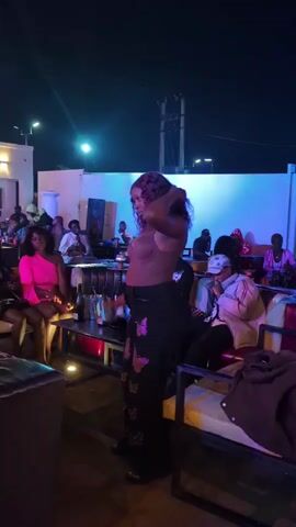 A woman with dark, curly hair and a tan top is dancing in front of a crowd of people. She is wearing dark pants with butterfly designs. The scene appears to be an outdoor venue at night, with colorful lighting illuminating the background. Several people are seated at tables and on benches, watching the dancer. Some individuals are holding drinks. The overall atmosphere seems to be festive and social.