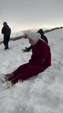 The video shows a young woman sledding down a snowy hill in a rural area. She is wearing a maroon suit, white shoes and a white hijab. Two other people are visible in the background, one standing and another crouched down. There are a few trees sparsely scattered across the snow-covered hillside. In the distance, a small town is visible. The sky is overcast. The woman slides down the hill on her back, enjoying the ride.