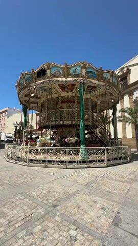 The video shows a full shot of an ornate carousel operating outdoors on a sunny day. The carousel is white with gold trim and decorated with colorful paintings of landscapes and sea scenes. Green fabric drapes hang from the roof. Several horses are visible, some occupied by people. The carousel is surrounded by a decorative white fence. The ground is paved with cobblestones. In the background, there are buildings, a palm tree, and a clear blue sky. The carousel is slowly rotating.