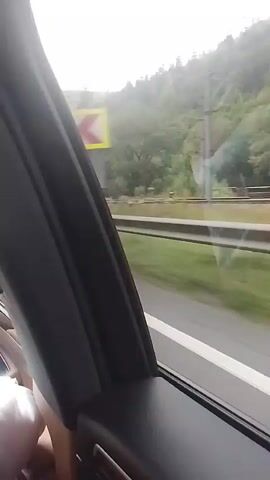 The video, filmed from a moving vehicle, shows a roadside scene in a mountainous, forested area. A guardrail runs along the side of the road, with a grassy verge and then a steep, tree-covered slope. The sky is overcast with some patches of blue. A traffic sign indicating a sharp left turn is visible early in the video. Later, a larger sign appears with the text "STATIUNEA AZUGA" (Azuga Resort) and several pictograms depicting activities such as skiing, a martini glass, and a snowboard. A black car is parked near the sign, and some logs are stacked to the right. The overall impression is of a scenic route leading to a mountain resort.