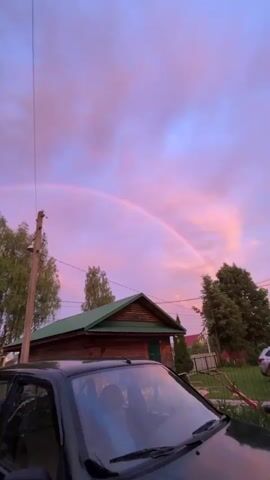 A video shows a faint rainbow against a pink and purple sunset sky. In the foreground, the top of a dark colored car is visible. Behind the car, a wooden house with a green roof stands. A fence and trees are also visible. The camera is panning slightly to the right.