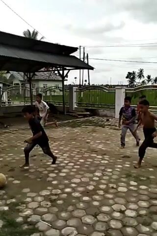 The video shows four barefoot young boys playing soccer in a yard. The yard is paved with a pattern of hexagonal stones. There is a roofed structure in the background with a metal fence around it, and some wooden construction materials are scattered inside the structure. One boy kicks the soccer ball, and it flies off-screen. Another boy is wearing a plaid shirt and red pants. A third boy is wearing a white t-shirt with the number "9" on it and shorts. The fourth boy is wearing a black t-shirt and denim shorts. The weather is overcast.