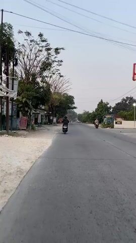 The video shows a wide paved road with a slight incline leading into the distance. On the left side of the road, there is a dirt shoulder lined with a few small shops or stalls, and trees. On the right side, there are more trees and some structures. The sky is overcast and light. Two motorcycles are visible on the road, both traveling away from the camera. The first motorcycle, a darker color, is closer to the center of the road. Further down the road, a second motorcycle, red in color, is also traveling away from the camera. The overall atmosphere is calm and suggests a rural or suburban setting.