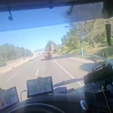The video shows a point-of-view shot from inside the cabin of a moving vehicle, likely a truck, traveling on a highway. The sky is clear and blue. The road is multi-laned with other vehicles visible.  There is a sign over the highway indicating “Lançon de Provence” with decorative towers on either side.  The dashboard of the vehicle is visible with two GPS devices, a steering wheel and other vehicle controls.  Trees line the side of the highway.  There is a sound of an air horn.