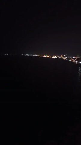 A night view of a city skyline by the water. The lights of the city are visible in the distance, reflecting on the water. The sky is dark, indicating it is nighttime. The foreground is dark, suggesting the camera is positioned some distance from the city. The water appears calm.