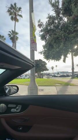 The video shows the view from the interior of a convertible car, with the top down. The car appears to be moving slowly along a road near a beach. Palm trees line the road, and a grassy area is visible between the road and a parking lot. Cars are parked in the parking lot, and the ocean is visible in the distance. The sky is partly cloudy, with the sun shining through the clouds. The car's interior features brown leather upholstery and metallic trim.