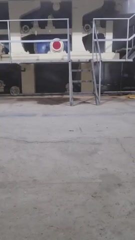 The camera pans right across a large, industrial machine in a warehouse. The machine is predominantly white and gray, with some black paneling. It has multiple levels and metal railing around the upper sections. A small set of metal stairs provides access to the upper level. The machine appears to be stationary. The floor is concrete and cracked, with some debris scattered around. The walls of the warehouse are corrugated metal. There are no people visible in the shot. The lighting is somewhat dim, contributing to the industrial atmosphere.