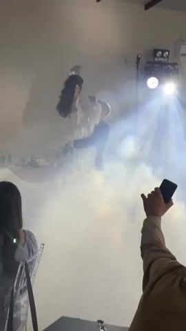 A wedding reception is underway. A bride in a white gown and a groom in a black suit are dancing in a cloud of smoke, illuminated by spotlights. The bride is being held up by the groom, her arms outstretched. The guests are watching the dance. One guest is filming the event on their phone. The room appears to be a large hall with exposed beams on the ceiling.
