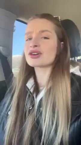 A woman with long blonde hair is sitting in a car. She is wearing a black leather jacket over a white top. She is looking directly at the camera and speaking. She briefly glances to the side, seemingly looking out the window. The car appears to be moving. There is a clear blue sky visible through the window. The woman is wearing makeup, including lipstick. The car's seatbelt is visible.