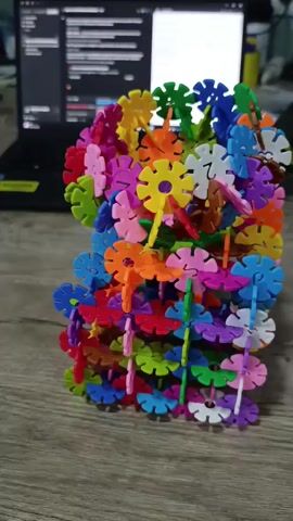 The video shows a close-up view of a colorful, abstract structure made of interlocking plastic pieces. The structure is predominantly cylindrical and appears to be a toy or a decorative item. The pieces are shaped like snowflakes or flowers and come in various bright colors including red, blue, green, yellow, orange, pink, and purple. The structure is placed on a wooden surface, likely a table, in front of a laptop. The background is blurred, but a portion of the laptop screen and some other objects are faintly visible. The lighting suggests an indoor setting. There are no people or animals visible in the video.