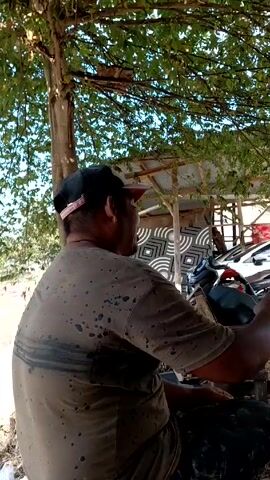 A man with a cap sits under a tree next to a river. The man is wearing a dark-colored t-shirt that has splotches and a thick stripe across the back. He is facing to his right, looking toward a building made of wood supports. A panel with black-and-white geometric designs leans against the side of the building. A motorcycle and a white car are parked nearby. The tree he is sitting under provides shade. The view from under the tree reveals the river.
