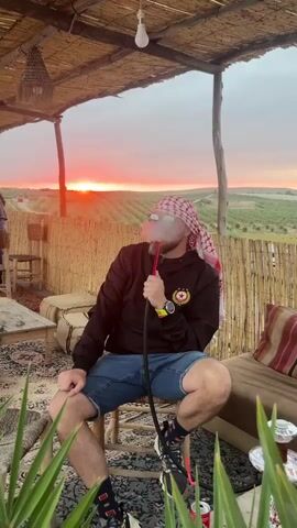A man wearing a black jacket with a red and gold emblem, denim shorts, sneakers, sunglasses, and a red and white keffiyeh is seated on a wooden stool. He is smoking a hookah, exhaling a cloud of white smoke from its hose. The setting is a rustic, semi-covered outdoor seating area, possibly a cafe or lounge, with a roof made of woven reeds and side walls of bamboo-like fencing. The floor is covered with a patterned rug, and low wooden tables, cushioned benches, and green leafy plants are visible in the foreground.

In the background, a dramatic sunset bathes the horizon in vibrant orange and red hues. The landscape beyond features rolling hills and what appear to be cultivated fields or olive groves. Another man is partially visible standing in the left background.

Notable objects include a simple lightbulb hanging from the roof, a woven basket-style light fixture, and a beverage can near the seated man's feet. The man appears relaxed, enjoying the serene atmosphere and the breathtaking view at sunset in Sidi Badhaj, Morocco.