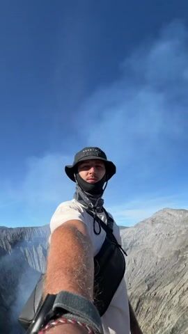 A first-person perspective video shows a person with a hat and bandana over their face standing at the edge of a large volcanic crater. The crater walls are steep and rocky, with plumes of smoke rising from the bottom. The sky is blue with some clouds. The person is holding the phone to take a selfie, and their arm and shoulder are visible in the foreground. The person is wearing a white t-shirt, a black hat, and a grey bandana. A black cross-body bag is visible. The video pans slightly, revealing more of the crater and the surrounding mountainous landscape.