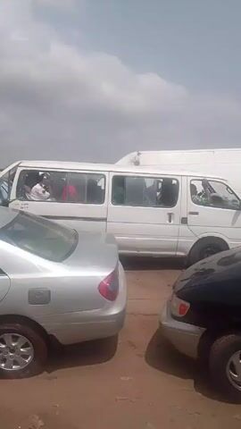 The video shows a street scene with a traffic jam. A black car with the logo "EXCELTECH" on the side is in the foreground. The video pans to show various other vehicles, including a white van. Several people are standing around the cars, some wearing uniformed outfits. There is trash and debris on the side of the road. A sign reads "BRAM-ACADEMY." The sky is overcast.