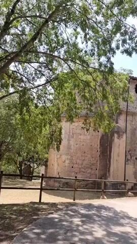 The video captures a sunny day in Sales de Llierca, Spain, with a view of an old stone building, likely a church, with a bell tower visible. Lush green trees frame the shot, their branches casting dappled shadows on a paved path. A wooden fence runs along the side of the path, separating it from a grassy area. The building itself is made of large, rough-hewn stones, with several small, arched windows along its upper wall. The sky is blue with a few wispy clouds, suggesting pleasant weather. The overall scene conveys a sense of peace and historical charm. There is no visible text in the video.