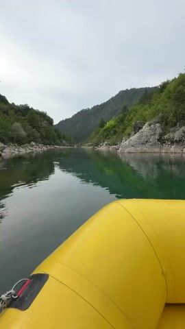This media file captures a group of people on a rafting trip on a river. The scene is set in a natural environment with lush green trees and rocky hills flanking the river. The water is a clear, emerald green, reflecting the overcast sky. The main subjects are five individuals seated in a yellow inflatable raft. They are all wearing life vests and red helmets, suggesting they are participating in an adventurous water activity.

In the foreground, a young woman is prominently featured, looking directly at the camera with a slight smile, indicating a positive and enjoyable experience. Behind her, four other individuals are seated in the raft. Two women in the middle are also wearing helmets and life vests, and one of them is holding a paddle. Towards the back of the raft, a man is visible, also wearing a helmet.

The raft itself is a bright yellow inflatable, with water droplets visible on its surface, hinting at the misty or wet conditions of the trip. The surrounding landscape suggests a mountainous or canyon-like terrain, with dense vegetation covering the slopes. The sky is overcast, creating a diffused light that is typical of a cloudy day.

The overall mood appears cheerful and adventurous, as evidenced by the smiles and the setting. The activity taking place is clearly white-water rafting or a similar form of river exploration. The presence of paddles and safety equipment reinforces this. The location cues, such as the type of vegetation and the rocky riverbanks, are consistent with a natural, somewhat wild environment, which is further supported by the provided location context of Džajići, Bosnia and Herzegovina. There is no visible text in the media file.