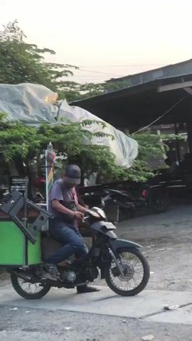 The video shows a person riding a motorcycle modified to carry a large, green cargo box. The rider, wearing a dark cap and a purple shirt, sits on the motorcycle, which appears to be moving. The motorcycle has a rack system attached behind the seat to support the cargo box. The cargo box itself is primarily green with some wooden accents and appears to be open at the top.

The scene is an outdoor environment with a dusty, unpaved road. In the background, there are some trees and structures that suggest a rural or semi-urban setting. One structure has a corrugated metal roof. The lighting suggests it is either early morning or late afternoon, with a warm, soft light. The overall atmosphere appears calm and ordinary, depicting a daily activity. There is no discernible text visible in the video. The activity captured is the transportation of goods or products using a customized motorcycle.