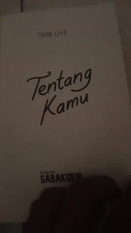 A person's hand is seen holding an open book. The book's cover displays the title "Tentang Kamu" in cursive font, with the author's name "TERE LIYE" above it and the publisher "PENERBIT SABAKGRIP" below. The pages are white with black text. The first page is mostly blank, with the title and author information. The subsequent pages show printed text in Indonesian. The camera is focused on the book, with the background being out of focus and appearing to be a tiled surface and a dark, indistinct area. The lighting is dim. The video consists of short clips that show the book being turned from one page to another.