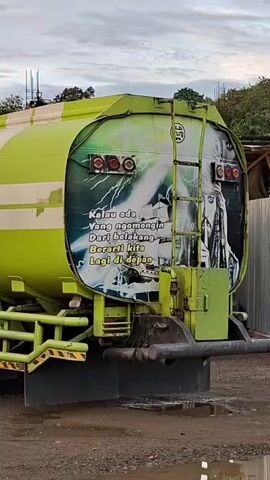 A bright green tanker truck, decorated with a white stripe and a central orange logo, is shown from its rear right side, parked on a dirt surface. The back of the truck features a painted scene of lightning and a person's silhouette, along with text in white. The truck then slowly moves forward to the left. The sky above is cloudy, and trees are visible in the background, along with what appears to be construction or industrial structures.