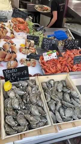 The video pans across a bustling seafood market stall. In the foreground, several wooden crates overflow with oysters. A bright yellow lemon rests on top of one crate. Behind the oysters, various types of shellfish are displayed on ice, including lobsters, crab claws, and large shrimp. Signs with French text and prices are visible, indicating different seafood selections. In the background, a person wearing a black shirt is partially visible, attending to the seafood display. The overall environment suggests a vibrant and fresh food market.