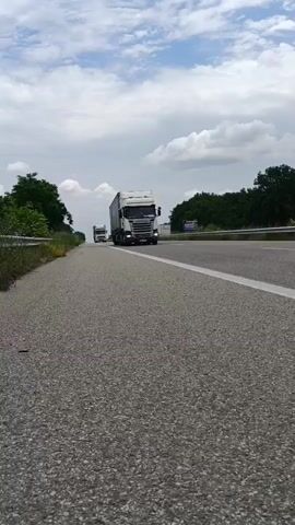 The video shows a low-angle shot of a highway with two semi-trucks driving on it. The truck in the foreground is white and has a large cargo trailer. The truck in the background is also white. The highway is paved and has a white line marking the lanes. There are trees and bushes on the side of the road. The sky is cloudy.