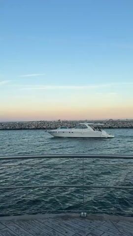 A white motor yacht is sailing on the sea with a stone breakwater in the background. The sky is a clear blue with a soft gradient of pink and orange towards the horizon, indicating it is either sunrise or sunset. The water is a deep blue-green with gentle waves. In the foreground, a metal railing with netting is visible, suggesting the vantage point is from a pier, balcony, or ship. The scene evokes a sense of calm and leisure, typical of a coastal setting. The time of day is suggested by the lighting and sky colors. There are no visible text elements in the media file.