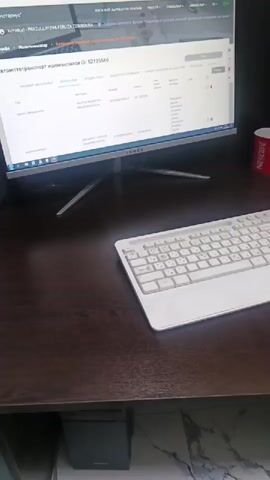 The video pans across a desk with a computer monitor, keyboard, and printer. On the monitor, a web page is displayed with Cyrillic text. A red mug with a logo is visible to the right of the monitor. A smartphone and some papers are next to the printer. A colorful mousepad with a white mouse is on the desk in front of the printer. The desk appears to be made of dark wood.