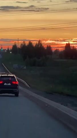A dark colored SUV is traveling on a paved road at dusk. The sky is a vibrant orange and yellow, indicating either sunrise or sunset. There are several other cars on the road ahead of the SUV, their headlights illuminated. Trees and other foliage line the right side of the road, and a guardrail is visible. Traffic lights are also present in the distance, some showing green. The overall scene depicts a typical evening commute on a highway, with the added beauty of a colorful sky.