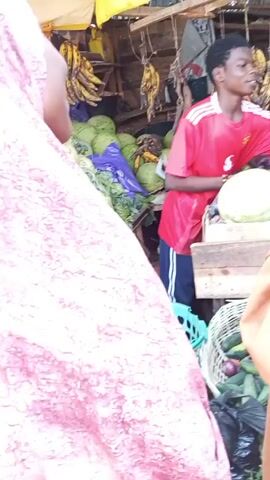 The video shows a bustling market scene with a young man in a red soccer jersey tending to his produce stand. He is smiling and interacting with customers, who are purchasing goods. The stand is filled with watermelons, plantains, and other fruits and vegetables. The customers are wearing colorful garments, indicating a vibrant and lively environment. The market appears to be outdoors, with a makeshift roof providing shade.