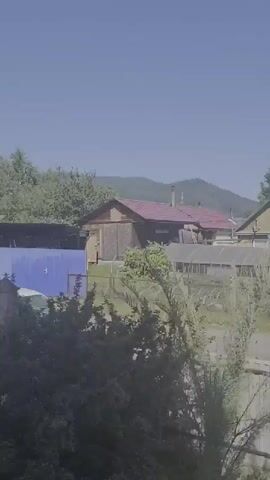 The video shows a rural landscape under a clear blue sky. In the foreground, the tops of trees and bushes are visible, with some dried grasses. In the middle ground, several buildings are present, including a wooden house with a red roof and a chimney, a blue-covered structure, and a greenhouse with a metal frame and transparent panels. Another house, painted green, has a satellite dish on its side. In the background, rolling green hills and mountains rise against the sky. The scene appears to be a residential area in a natural setting.