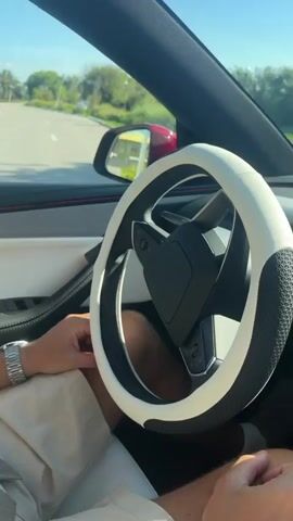 The video shows a person's hands on the steering wheel of a car, with the car's dashboard and a large touchscreen display visible. The steering wheel has a white and black cover. The touchscreen displays a navigation map and driving information, including speed and distance. The person's legs are also visible, and they are wearing shorts. Outside the car, trees and buildings can be seen, suggesting an outdoor setting. The car appears to be in motion, as indicated by the road and surroundings.