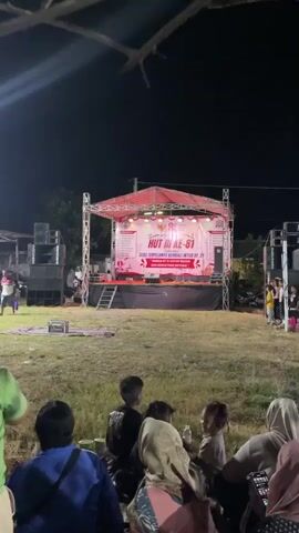 A nighttime outdoor scene shows a raised stage decorated with banners and lights. A crowd of people, including adults and children, sits on the grass watching the stage. Speakers are set up on either side of the stage.