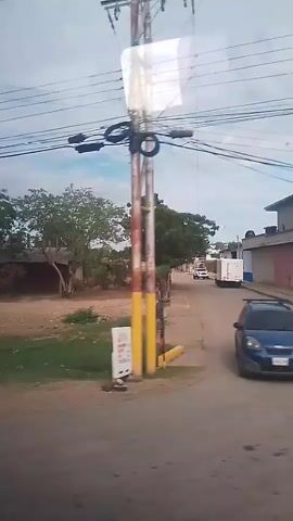 A handheld video, filmed from a moving vehicle, shows a rural or semi-rural area. Power lines are visible overhead. In the foreground, a dirt and grassy area with a few rocks is visible. In the middle ground, there are several buildings, including a small, weathered structure with a reddish-brown roof and walls, and another building with a similar roof. Trees and bushes are scattered throughout the scene. In the background, there is a white truck and a white cargo trailer. The sky is overcast.