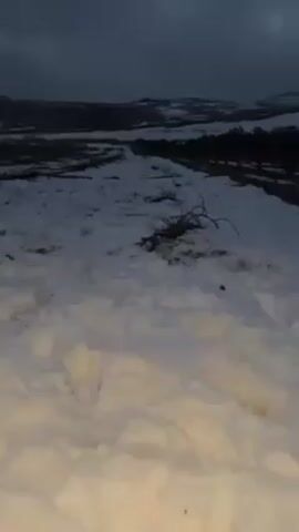 The video starts with a view of a snow-covered landscape under a dark, overcast sky. There are some small shrubs sticking out of the snow. In the distance, there appear to be rolling hills. Suddenly, a cloud of what appears to be smoke or fog is seen up close, obscuring the view. The smoke/fog is white and gray.