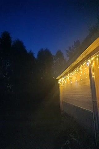 This is a short video clip, showing a wooden structure, possibly a shed or a house, at dusk. The structure is decorated with a string of yellow, icicle-shaped lights along its roofline. The lights are illuminated, casting a warm glow. To the left of the structure, silhouetted trees and foliage are visible against a darkening blue sky. There appears to be some fog or mist in the air, particularly between the trees and the structure, which is catching the light from the string lights. The ground in front of the structure is dark and appears to be grass or dirt. There are no people or animals visible in the video. The overall atmosphere is calm and serene, suggesting a quiet evening setting.