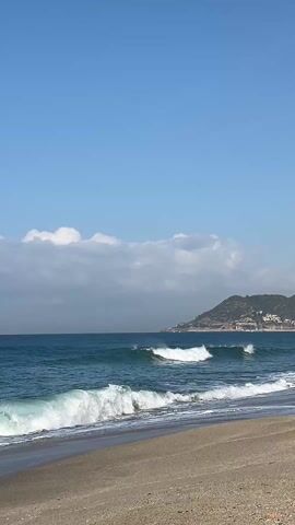 The video shows a beach on a sunny day with waves crashing onto the shore. The water is a deep blue-green color. The sand is a light tan color, and there are some small pebbles mixed in. The sky is blue with some white clouds. The video is shot from a static position.