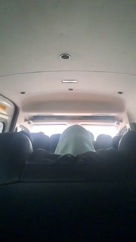 The video captures the interior of a passenger vehicle, likely a van or minibus, viewed from a rear seat looking forward. Several passengers are visible from behind, their dark silhouettes contrasting with the bright light streaming through the front and side windows. One passenger in the center, directly in front of the camera's perspective, is wearing a light-colored head covering. The vehicle's ceiling is light-colored, featuring multiple recessed lights and air vents. The seats are upholstered in a dark material, likely black. The scene appears to be taking place during the daytime, indicated by the strong natural light entering the vehicle. The vehicle seems to be in motion, suggested by the slight camera movement and ambient sound, though no specific activity other than traveling is visible. No text is discernible in the frame. The location is Ujong Duabelas, Indonesia.