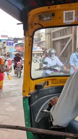 The video shows a street scene, likely in India, from the perspective of someone inside an auto-rickshaw. The camera is pointed forward and slightly to the right. In the foreground, the yellow and green interior of the auto-rickshaw is visible, including a steering wheel and a driver's hand. On the street, there are several people walking and riding on motorcycles and auto-rickshaws. A woman in a bright pink and yellow sari is walking away from the camera on the left side of the street. Further down the street, there are buildings and a temple with a white dome and orange trim. The sky is overcast.