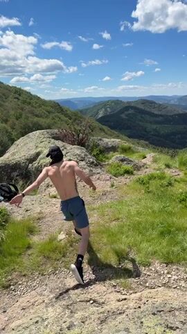 A shirtless man wearing shorts and a bandana performs a backflip on a grassy hilltop overlooking a valley. He is positioned on a rocky outcrop with a backpack to his left. The sky is blue with scattered clouds.