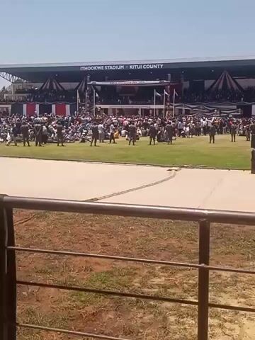 The video shows a large crowd gathered at Ithookwe Stadium in Kitui County. People are standing and sitting in the stadium, with uniformed personnel present. The camera moves around to show different sections of the crowd. Some people are standing near a railing overlooking the field. There's a flag present and the video ends.