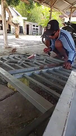 A person wearing a blue shirt and a black cap crouches and welds a metal frame. They are holding a welding torch and a metal rod. The frame is made of silver metal bars and is lying on the ground. There is a motorcycle parked behind the person, and a building in the background. The scene appears to be outdoors, under a corrugated metal roof.