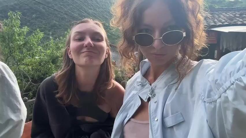 Two young women are posing for a selfie. The woman on the left is making a peace sign with her fingers, and later puts up all five fingers. She is wearing a dark gray top and a black bag is slung over her shoulder. The woman on the right is wearing sunglasses and a light blue jacket over a pink top. She smiles, and then adopts a more serious expression. The background shows lush green trees, a building with a tiled roof, and a woman in traditional clothing. The scene seems to be a mountainous or rural area.