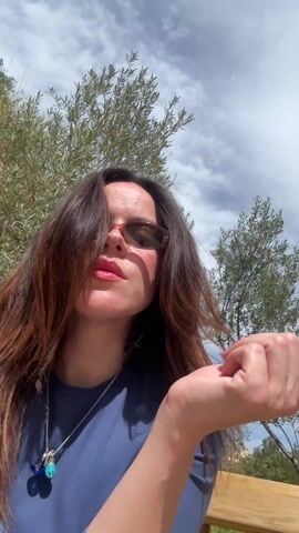 A woman in sunglasses and a blue shirt takes a selfie in a park on a sunny day. She sits on a wooden bench surrounded by greenery and yellow flowers. In the background, there's a wooden fence and residential buildings. A small dog walks along the other side of the fence in one shot.