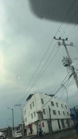 The video, shot from inside a car on a cloudy day, shows a white, multi-story building with multiple windows, appearing to be located on a street corner. A small shrine or religious image is visible on the building’s facade. In the foreground, a person on a motorcycle is parked near the building. A power pole with numerous wires extends into the sky, partially obscuring the view. The windshield wipers activate momentarily, clearing raindrops from the glass. A streetlamp and a sign for "Evergreen" are also visible in the scene.