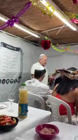 The scene takes place in a decorated Sukkah. An older man in a white shirt and kippah holds a platter and smiles, presumably leading a religious service. A younger woman with blonde hair is serving food, holding a tray with what appears to be flatbread. Other people are seated around a table covered with a white tablecloth. On the table are various dishes, including salads, a bottle of orange juice, and drinks. The walls of the Sukkah are decorated with a large poster in Hebrew and various festive decorations.
