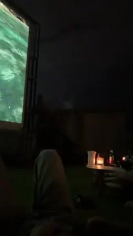 The video shows a person sitting on a lounge chair, with a projected image displayed on a screen in front of them. The person is wearing light-colored shorts. In the initial frames, the projected image displays abstract patterns. As the video progresses, the image transitions to a scene showing a woman sitting on the steps of a building adorned with lush greenery and flowers. Adjacent to the chair, on a small table, are two glasses with straws and a lit candle. The surrounding environment appears to be outdoors, possibly a rooftop or patio, given the dimly lit sky visible in the background.