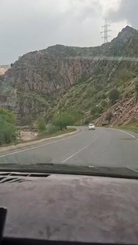 The video shows a car driving on a winding road through a mountainous area. The road is paved and has a white line on the edge. There are rocky cliffs and some greenery on either side of the road. There is a river flowing near the road. In the distance, there is an electricity pylon on top of a hill. The sky is overcast. The view is from the driver's perspective. A pack of tissues sits on the dashboard.