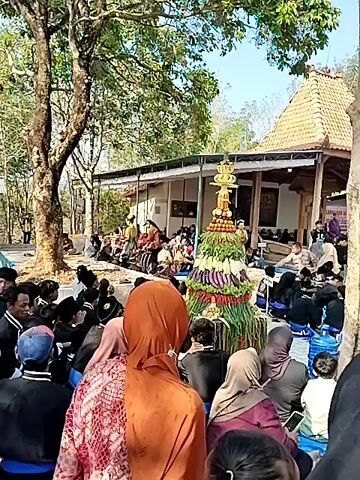 A large group of people are gathered outdoors in what appears to be a village or rural setting. In the foreground, a tall, conical structure decorated with fruits and vegetables is prominently displayed. Many individuals, some wearing headscarves, are seated on the ground or on low benches, facing the structure and a building in the background. The building has a traditional tiled roof and appears to be a place of worship or a community hall. Several trees surround the gathering, and the ground is mostly dirt or gravel. The overall atmosphere suggests a communal event or ceremony.
